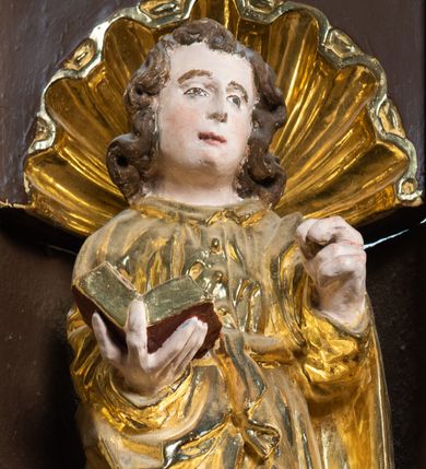 Zdjęcie nr 19: Ambona drewniana, przyścienna, wisząca, dostawiona do północnej ściany nawy ze schodkami biegnącymi wzdłuż ściany. Kosz ambony jest sześcioboczny, z niewielkim parapetem. Jego boki zostały podzielone na pola stojącymi na wysokich cokołach pięcioma kolumienkami o korynckich kapitelach, dźwigających profilowane belkowanie. W polach prostokątne płyciny z konchowymi wnękami ozdobionymi muszlami z figurami ewangelistów. 
We wnękach rzeźby z przedstawieniami ewangelistów i Chrystusa. Figury drewniane pełnoplastyczne, wydrążone od tyłu, polichromowane, złocone i srebrzone, ustawione na niewielkich prostokątnych podstawach, od lewej: św. Łukasz wsparty prawą nogą na głowie byka, św. Marek wsparty prawą nogą na głowie lwa, św. Jan z orłem przy prawej nodze, Chrystus Salvator Mundi, św. Mateusz z aniołem przy prawej nodze. Ewangeliści trzymają w prawych dłoniach otwarte księgi, z wyjątkiem św. Mateusza, trzymającego kodeks w lewej ręce. Dłonie ewangelistów nieproporcjonalnie duże. Szaty bogato pofałdowane, głęboko cięte. 
Podstawa kosza bogato profilowana, ozdobiona podwieszoną dekoracyjną szyszką. Schodki ograniczone parapetem z trzema romboidalnymi płycinami w złoconych profilowanych obramieniach. Na filarze przy wejściu na schodki złocona kula z szyszką. Zaplecek w kształcie stojącego prostokąta, w górnej części ujęty zwężającymi się ku dołowi pilastrami wypełnionymi cekinami, zwieńczonymi rombowymi wypustkami i korynckimi kapitelami, które podtrzymują przełamujące się nad nimi belkowanie. Pośrodku zaplecka w prostokątnym obramieniu zamkniętym nadwieszonym łukiem półkolistym płaskorzeźbione popiersie Boga Ojca wyłaniającego się z obłoków. Prawa ręka Boga wzniesiona w geście błogosławieństwa, lewa oparta na globie ziemskim zwieńczonym krzyżem. Tłem dla postaci Boga Ojca jest rozwiana poła płaszcza układająca się niemal półkoliście. Baldachim ośmioboczny. W profilowanym podniebiu, ozdobionym pasem ornamentu małżowinowo-chrząstkowego i profilowanymi wisiorami, gołębica Ducha Świętego na tle glorii utworzonej z prostych promieni o równej długości. Baldachim ozdobiony zamkniętą koroną, którą tworzy pięć esownic pokrytych pasami ornamentu małżowinowo-chrząstkowego. W zwieńczeniu pełnoplastyczna rzeźba przedstawiająca św. Jana Chrzciciela. Figura drewniana, wydrążona z tyłu, polichromowana, złocona i srebrzona, ustawiona na niewielkiej wielobocznej profilowanej podstawie. Święty w prawej ręce trzyma wysoki krzyż, w lewej księgę, na której spoczywa Baranek Boży. Struktura ambony ciemnobrązowa. Kolumny, kapitele, pilastry w zaplecku korona baldachimu, profilowania i ornamenty złocone, z wyjątkiem srebrzonej ramy w zaplecku i gołębicy Ducha Świętego.