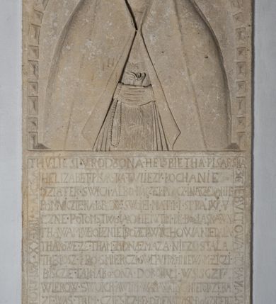 Zdjęcie nr 5: Nagrobek w formie płaskorzeźbionej tablicy o kształcie stojącego prostokąta. W jego górnej części znajduje się wyobrażenie Elżbiety z Giebułtowskich Pisarskiej przedstawionej frontalnie, do pasa. Kobieta jest obramiona półkolistą arkadą utworzoną z kaboszonów. Po bokach arkady dwa ozdobione rollwerkami kartusze z godłami herbów Bersten (po lewej stronie) i Jelita (po prawej stronie). Wyłaniająca się spod szat twarz zmarłej o kształcie sercowatym, z pełnymi policzkami z dużymi zamkniętymi oczami, z prostym, wydatnym nosem, wąskimi ustami i wyraźnie zaznaczonym podbródkiem. Zmarła jest ubrana we wdowi welon (rańtuch) spięty pod brodą i rozchylający się od środka na boki. Gładki welon zdobi jedna rurkowata fałda biegnąca na całej jego długości. Głowę nakrywa czepek, a szyję zasłania podwika. Spódnica spięta pasem z ozdobnymi klamrami, układana w drobne rurkowate fałdy. Ręce złożone, w nich chustka, przez prawą rękę przewieszony różaniec. W dolnej partii nagrobka inskrypcja kuta majuskułą. Treść inskrypcji w otoku bordiury i w polu. Inskrypcja w otoku: Inskrypcja na bordiurze &quot;THU. LIESI. URODZONA. HELZBIETHA. PISARSKA / Z GIEBVLTOWA. KTORA MAIANCZ WIEKU / SWEGO. LIATTH. LVI. S. STHEM. SZWIATHEM. / ROZL/ONCZONA. W ROKV. 1576. XX. DNIA MARCEA.&quot; Inskrypcja w polu: &quot;HELIZABET. PISARSKA. TV LIEZY. KOCHANIE. / DZIATEK. SWICH. ALBO. RACZEI. BACZ. I NARZEKANIE / POMNICZIE. NA. POBOŻNE. SWOIEI. MATKI SPRAWY/CZNE POTOMSTWO IAKO I IEI W TIM. BIŁ BOG. ŁASKAWY / THA WAM W BOIZNIE I BOZEJ. WYCHOWANIE DAŁA / THAKOWE. ZE. THAM. ZADNA. ZMAZA. NIEZOSTAŁA / THEGOSZ. I. PO. SMIERCZI. SWEI. VPRZYIMIE WAM. ZICZI. / BISZCIE. TAK. IAKO. ONA. DOKONALI. WSISCZI. / WIEKOW. SWOICH. A W TIM. WAM WĄTPICZ NIEPOTRZEBA. / ZE. WAS. THIM. CZIESICZ. BENDZIE. NAIWISI. BOG. Z NIEBA. / I. TIM. WAM. TO. ZAPLACZI. ZE. DNIA. OSTATNIEGO. / STANIECZIE. NAPRAWICZI. Z WIBRANEMI. IEGO. / BARBARA TOMASOWSKA. TEN. KAMIEŃ. POSTAWICZ. DAŁA&quot;. 


