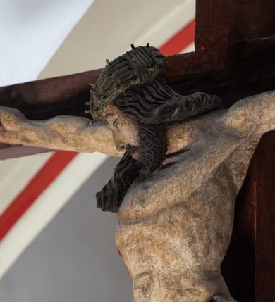 Zdjęcie nr 4: Rzeźba drewniana, pełnoplastyczna, polichromowana przedstawia Chrystusa Ukrzyżowanego i jest umieszczona na profilowanej belce tęczowej wspartej na kroksztynach. Chrystus jest ukazany frontalnie, w typie Christo morto, przybity do krzyża trzema gwoździami. Jego ramiona są silnie wyprężone, rozłożone horyzontalnie. Głowa w ciężkiej koronie cierniowej, opada na prawe ramię. Twarz ma owalną, oczy zamknięte, nos wydatny, usta lekko rozchylone. Twarz okala bujny, ciemny zarost. Długie ciemne włosy ułożone w pukle opadają po prawej stornie na ramie, po lewej na plecy Chrystusa. Korpus z zaznaczoną klatką piersiową, wyraźnym przewężeniem w talii, w prawym boku widoczna krwawiąca rana. Perizonium na biodrach jest krótkie, przewiązane, z końcem opadającym wzdłuż lewego boku. Nogi lekko ugięte w kolanach, stopy skrzyżowane, prawa założona na lewą, przebite jednym gwoździem. Na belce krzyża, nad głową Chrystusa titulus w kształcie banderoli. Ciało Chrystusa w kolorze naturalnym. Perizonium jest w kolorze perłowo-szarym, z błękitną podszewką.