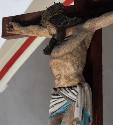 Zdjęcie nr 5: Rzeźba drewniana, pełnoplastyczna, polichromowana przedstawia Chrystusa Ukrzyżowanego i jest umieszczona na profilowanej belce tęczowej wspartej na kroksztynach. Chrystus jest ukazany frontalnie, w typie Christo morto, przybity do krzyża trzema gwoździami. Jego ramiona są silnie wyprężone, rozłożone horyzontalnie. Głowa w ciężkiej koronie cierniowej, opada na prawe ramię. Twarz ma owalną, oczy zamknięte, nos wydatny, usta lekko rozchylone. Twarz okala bujny, ciemny zarost. Długie ciemne włosy ułożone w pukle opadają po prawej stornie na ramie, po lewej na plecy Chrystusa. Korpus z zaznaczoną klatką piersiową, wyraźnym przewężeniem w talii, w prawym boku widoczna krwawiąca rana. Perizonium na biodrach jest krótkie, przewiązane, z końcem opadającym wzdłuż lewego boku. Nogi lekko ugięte w kolanach, stopy skrzyżowane, prawa założona na lewą, przebite jednym gwoździem. Na belce krzyża, nad głową Chrystusa titulus w kształcie banderoli. Ciało Chrystusa w kolorze naturalnym. Perizonium jest w kolorze perłowo-szarym, z błękitną podszewką.