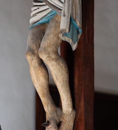 Zdjęcie nr 7: Rzeźba drewniana, pełnoplastyczna, polichromowana przedstawia Chrystusa Ukrzyżowanego i jest umieszczona na profilowanej belce tęczowej wspartej na kroksztynach. Chrystus jest ukazany frontalnie, w typie Christo morto, przybity do krzyża trzema gwoździami. Jego ramiona są silnie wyprężone, rozłożone horyzontalnie. Głowa w ciężkiej koronie cierniowej, opada na prawe ramię. Twarz ma owalną, oczy zamknięte, nos wydatny, usta lekko rozchylone. Twarz okala bujny, ciemny zarost. Długie ciemne włosy ułożone w pukle opadają po prawej stornie na ramie, po lewej na plecy Chrystusa. Korpus z zaznaczoną klatką piersiową, wyraźnym przewężeniem w talii, w prawym boku widoczna krwawiąca rana. Perizonium na biodrach jest krótkie, przewiązane, z końcem opadającym wzdłuż lewego boku. Nogi lekko ugięte w kolanach, stopy skrzyżowane, prawa założona na lewą, przebite jednym gwoździem. Na belce krzyża, nad głową Chrystusa titulus w kształcie banderoli. Ciało Chrystusa w kolorze naturalnym. Perizonium jest w kolorze perłowo-szarym, z błękitną podszewką.