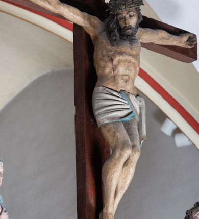 Zdjęcie nr 6: Rzeźba drewniana, pełnoplastyczna, polichromowana przedstawia Chrystusa Ukrzyżowanego i jest umieszczona na profilowanej belce tęczowej wspartej na kroksztynach. Chrystus jest ukazany frontalnie, w typie Christo morto, przybity do krzyża trzema gwoździami. Jego ramiona są silnie wyprężone, rozłożone horyzontalnie. Głowa w ciężkiej koronie cierniowej, opada na prawe ramię. Twarz ma owalną, oczy zamknięte, nos wydatny, usta lekko rozchylone. Twarz okala bujny, ciemny zarost. Długie ciemne włosy ułożone w pukle opadają po prawej stornie na ramie, po lewej na plecy Chrystusa. Korpus z zaznaczoną klatką piersiową, wyraźnym przewężeniem w talii, w prawym boku widoczna krwawiąca rana. Perizonium na biodrach jest krótkie, przewiązane, z końcem opadającym wzdłuż lewego boku. Nogi lekko ugięte w kolanach, stopy skrzyżowane, prawa założona na lewą, przebite jednym gwoździem. Na belce krzyża, nad głową Chrystusa titulus w kształcie banderoli. Ciało Chrystusa w kolorze naturalnym. Perizonium jest w kolorze perłowo-szarym, z błękitną podszewką.