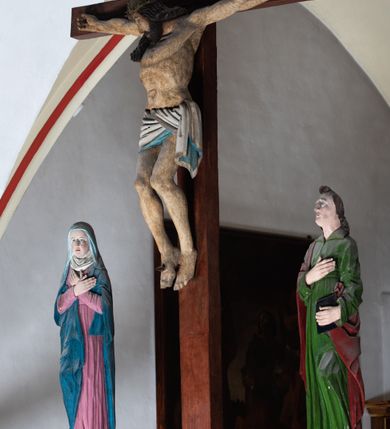 Zdjęcie nr 8: Rzeźba drewniana, pełnoplastyczna, polichromowana przedstawia Chrystusa Ukrzyżowanego i jest umieszczona na profilowanej belce tęczowej wspartej na kroksztynach. Chrystus jest ukazany frontalnie, w typie Christo morto, przybity do krzyża trzema gwoździami. Jego ramiona są silnie wyprężone, rozłożone horyzontalnie. Głowa w ciężkiej koronie cierniowej, opada na prawe ramię. Twarz ma owalną, oczy zamknięte, nos wydatny, usta lekko rozchylone. Twarz okala bujny, ciemny zarost. Długie ciemne włosy ułożone w pukle opadają po prawej stornie na ramie, po lewej na plecy Chrystusa. Korpus z zaznaczoną klatką piersiową, wyraźnym przewężeniem w talii, w prawym boku widoczna krwawiąca rana. Perizonium na biodrach jest krótkie, przewiązane, z końcem opadającym wzdłuż lewego boku. Nogi lekko ugięte w kolanach, stopy skrzyżowane, prawa założona na lewą, przebite jednym gwoździem. Na belce krzyża, nad głową Chrystusa titulus w kształcie banderoli. Ciało Chrystusa w kolorze naturalnym. Perizonium jest w kolorze perłowo-szarym, z błękitną podszewką.