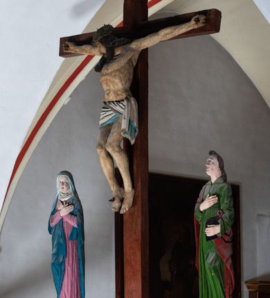 Zdjęcie nr 9: Rzeźba drewniana, pełnoplastyczna, polichromowana przedstawia Chrystusa Ukrzyżowanego i jest umieszczona na profilowanej belce tęczowej wspartej na kroksztynach. Chrystus jest ukazany frontalnie, w typie Christo morto, przybity do krzyża trzema gwoździami. Jego ramiona są silnie wyprężone, rozłożone horyzontalnie. Głowa w ciężkiej koronie cierniowej, opada na prawe ramię. Twarz ma owalną, oczy zamknięte, nos wydatny, usta lekko rozchylone. Twarz okala bujny, ciemny zarost. Długie ciemne włosy ułożone w pukle opadają po prawej stornie na ramie, po lewej na plecy Chrystusa. Korpus z zaznaczoną klatką piersiową, wyraźnym przewężeniem w talii, w prawym boku widoczna krwawiąca rana. Perizonium na biodrach jest krótkie, przewiązane, z końcem opadającym wzdłuż lewego boku. Nogi lekko ugięte w kolanach, stopy skrzyżowane, prawa założona na lewą, przebite jednym gwoździem. Na belce krzyża, nad głową Chrystusa titulus w kształcie banderoli. Ciało Chrystusa w kolorze naturalnym. Perizonium jest w kolorze perłowo-szarym, z błękitną podszewką.