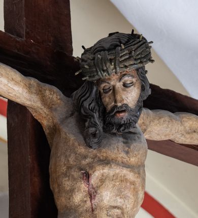 Zdjęcie nr 1: Rzeźba drewniana, pełnoplastyczna, polichromowana przedstawia Chrystusa Ukrzyżowanego i jest umieszczona na profilowanej belce tęczowej wspartej na kroksztynach. Chrystus jest ukazany frontalnie, w typie Christo morto, przybity do krzyża trzema gwoździami. Jego ramiona są silnie wyprężone, rozłożone horyzontalnie. Głowa w ciężkiej koronie cierniowej, opada na prawe ramię. Twarz ma owalną, oczy zamknięte, nos wydatny, usta lekko rozchylone. Twarz okala bujny, ciemny zarost. Długie ciemne włosy ułożone w pukle opadają po prawej stornie na ramie, po lewej na plecy Chrystusa. Korpus z zaznaczoną klatką piersiową, wyraźnym przewężeniem w talii, w prawym boku widoczna krwawiąca rana. Perizonium na biodrach jest krótkie, przewiązane, z końcem opadającym wzdłuż lewego boku. Nogi lekko ugięte w kolanach, stopy skrzyżowane, prawa założona na lewą, przebite jednym gwoździem. Na belce krzyża, nad głową Chrystusa titulus w kształcie banderoli. Ciało Chrystusa w kolorze naturalnym. Perizonium jest w kolorze perłowo-szarym, z błękitną podszewką.
