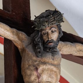 Zdjęcie nr 1: Rzeźba drewniana, pełnoplastyczna, polichromowana przedstawia Chrystusa Ukrzyżowanego i jest umieszczona na profilowanej belce tęczowej wspartej na kroksztynach. Chrystus jest ukazany frontalnie, w typie Christo morto, przybity do krzyża trzema gwoździami. Jego ramiona są silnie wyprężone, rozłożone horyzontalnie. Głowa w ciężkiej koronie cierniowej, opada na prawe ramię. Twarz ma owalną, oczy zamknięte, nos wydatny, usta lekko rozchylone. Twarz okala bujny, ciemny zarost. Długie ciemne włosy ułożone w pukle opadają po prawej stornie na ramie, po lewej na plecy Chrystusa. Korpus z zaznaczoną klatką piersiową, wyraźnym przewężeniem w talii, w prawym boku widoczna krwawiąca rana. Perizonium na biodrach jest krótkie, przewiązane, z końcem opadającym wzdłuż lewego boku. Nogi lekko ugięte w kolanach, stopy skrzyżowane, prawa założona na lewą, przebite jednym gwoździem. Na belce krzyża, nad głową Chrystusa titulus w kształcie banderoli. Ciało Chrystusa w kolorze naturalnym. Perizonium jest w kolorze perłowo-szarym, z błękitną podszewką.