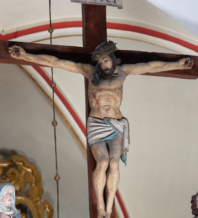 Zdjęcie nr 11: Rzeźba drewniana, pełnoplastyczna, polichromowana przedstawia Chrystusa Ukrzyżowanego i jest umieszczona na profilowanej belce tęczowej wspartej na kroksztynach. Chrystus jest ukazany frontalnie, w typie Christo morto, przybity do krzyża trzema gwoździami. Jego ramiona są silnie wyprężone, rozłożone horyzontalnie. Głowa w ciężkiej koronie cierniowej, opada na prawe ramię. Twarz ma owalną, oczy zamknięte, nos wydatny, usta lekko rozchylone. Twarz okala bujny, ciemny zarost. Długie ciemne włosy ułożone w pukle opadają po prawej stornie na ramie, po lewej na plecy Chrystusa. Korpus z zaznaczoną klatką piersiową, wyraźnym przewężeniem w talii, w prawym boku widoczna krwawiąca rana. Perizonium na biodrach jest krótkie, przewiązane, z końcem opadającym wzdłuż lewego boku. Nogi lekko ugięte w kolanach, stopy skrzyżowane, prawa założona na lewą, przebite jednym gwoździem. Na belce krzyża, nad głową Chrystusa titulus w kształcie banderoli. Ciało Chrystusa w kolorze naturalnym. Perizonium jest w kolorze perłowo-szarym, z błękitną podszewką.