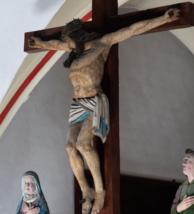 Zdjęcie nr 10: Rzeźba drewniana, pełnoplastyczna, polichromowana przedstawia Chrystusa Ukrzyżowanego i jest umieszczona na profilowanej belce tęczowej wspartej na kroksztynach. Chrystus jest ukazany frontalnie, w typie Christo morto, przybity do krzyża trzema gwoździami. Jego ramiona są silnie wyprężone, rozłożone horyzontalnie. Głowa w ciężkiej koronie cierniowej, opada na prawe ramię. Twarz ma owalną, oczy zamknięte, nos wydatny, usta lekko rozchylone. Twarz okala bujny, ciemny zarost. Długie ciemne włosy ułożone w pukle opadają po prawej stornie na ramie, po lewej na plecy Chrystusa. Korpus z zaznaczoną klatką piersiową, wyraźnym przewężeniem w talii, w prawym boku widoczna krwawiąca rana. Perizonium na biodrach jest krótkie, przewiązane, z końcem opadającym wzdłuż lewego boku. Nogi lekko ugięte w kolanach, stopy skrzyżowane, prawa założona na lewą, przebite jednym gwoździem. Na belce krzyża, nad głową Chrystusa titulus w kształcie banderoli. Ciało Chrystusa w kolorze naturalnym. Perizonium jest w kolorze perłowo-szarym, z błękitną podszewką.