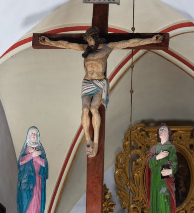 Zdjęcie nr 3: Rzeźba drewniana, pełnoplastyczna, polichromowana przedstawia Chrystusa Ukrzyżowanego i jest umieszczona na profilowanej belce tęczowej wspartej na kroksztynach. Chrystus jest ukazany frontalnie, w typie Christo morto, przybity do krzyża trzema gwoździami. Jego ramiona są silnie wyprężone, rozłożone horyzontalnie. Głowa w ciężkiej koronie cierniowej, opada na prawe ramię. Twarz ma owalną, oczy zamknięte, nos wydatny, usta lekko rozchylone. Twarz okala bujny, ciemny zarost. Długie ciemne włosy ułożone w pukle opadają po prawej stornie na ramie, po lewej na plecy Chrystusa. Korpus z zaznaczoną klatką piersiową, wyraźnym przewężeniem w talii, w prawym boku widoczna krwawiąca rana. Perizonium na biodrach jest krótkie, przewiązane, z końcem opadającym wzdłuż lewego boku. Nogi lekko ugięte w kolanach, stopy skrzyżowane, prawa założona na lewą, przebite jednym gwoździem. Na belce krzyża, nad głową Chrystusa titulus w kształcie banderoli. Ciało Chrystusa w kolorze naturalnym. Perizonium jest w kolorze perłowo-szarym, z błękitną podszewką.