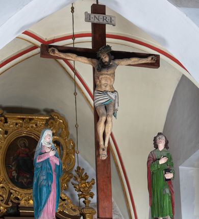 Zdjęcie nr 2: Rzeźba drewniana, pełnoplastyczna, polichromowana przedstawia Chrystusa Ukrzyżowanego i jest umieszczona na profilowanej belce tęczowej wspartej na kroksztynach. Chrystus jest ukazany frontalnie, w typie Christo morto, przybity do krzyża trzema gwoździami. Jego ramiona są silnie wyprężone, rozłożone horyzontalnie. Głowa w ciężkiej koronie cierniowej, opada na prawe ramię. Twarz ma owalną, oczy zamknięte, nos wydatny, usta lekko rozchylone. Twarz okala bujny, ciemny zarost. Długie ciemne włosy ułożone w pukle opadają po prawej stornie na ramie, po lewej na plecy Chrystusa. Korpus z zaznaczoną klatką piersiową, wyraźnym przewężeniem w talii, w prawym boku widoczna krwawiąca rana. Perizonium na biodrach jest krótkie, przewiązane, z końcem opadającym wzdłuż lewego boku. Nogi lekko ugięte w kolanach, stopy skrzyżowane, prawa założona na lewą, przebite jednym gwoździem. Na belce krzyża, nad głową Chrystusa titulus w kształcie banderoli. Ciało Chrystusa w kolorze naturalnym. Perizonium jest w kolorze perłowo-szarym, z błękitną podszewką.