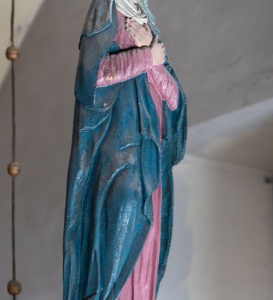 Zdjęcie nr 2: Rzeźba drewniana, pełnoplastyczna, polichromowana przedstawia Matkę Boską stojącą na niewielkim postumencie w kształcie zbliżonym do połowy sfery. Matka Boska ukazana w pozycji stojącej, frontalnie, w lekkim kontrapoście, z ciałem lekko zwróconym w kierunku krzyża, z rękoma skrzyżowanymi na piersi. Jej głowa jest nakryta welonem. Twarz ma owalną, oczy z opadającymi kącikami, nos prosty, a usta małe. Maria ubrana w długą suknię w kolorze zgaszonego różu przepasaną w stanie oraz niebieski płaszcz narzucony na ramiona  i opływający sylwetkę, na szyi udrapowana chusta. Fałdy szaty pionowe, płytkie, układające się miękko.