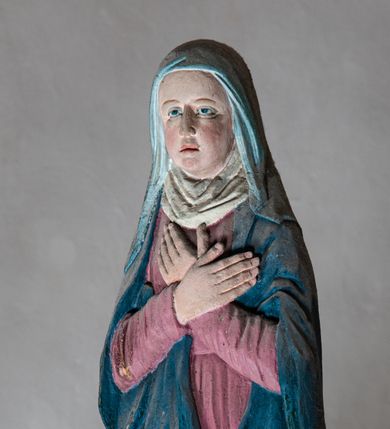 Zdjęcie nr 1: Rzeźba drewniana, pełnoplastyczna, polichromowana przedstawia Matkę Boską stojącą na niewielkim postumencie w kształcie zbliżonym do połowy sfery. Matka Boska ukazana w pozycji stojącej, frontalnie, w lekkim kontrapoście, z ciałem lekko zwróconym w kierunku krzyża, z rękoma skrzyżowanymi na piersi. Jej głowa jest nakryta welonem. Twarz ma owalną, oczy z opadającymi kącikami, nos prosty, a usta małe. Maria ubrana w długą suknię w kolorze zgaszonego różu przepasaną w stanie oraz niebieski płaszcz narzucony na ramiona  i opływający sylwetkę, na szyi udrapowana chusta. Fałdy szaty pionowe, płytkie, układające się miękko.