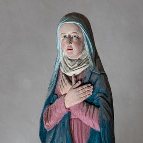 Zdjęcie nr 1: Rzeźba drewniana, pełnoplastyczna, polichromowana przedstawia Matkę Boską stojącą na niewielkim postumencie w kształcie zbliżonym do połowy sfery. Matka Boska ukazana w pozycji stojącej, frontalnie, w lekkim kontrapoście, z ciałem lekko zwróconym w kierunku krzyża, z rękoma skrzyżowanymi na piersi. Jej głowa jest nakryta welonem. Twarz ma owalną, oczy z opadającymi kącikami, nos prosty, a usta małe. Maria ubrana w długą suknię w kolorze zgaszonego różu przepasaną w stanie oraz niebieski płaszcz narzucony na ramiona  i opływający sylwetkę, na szyi udrapowana chusta. Fałdy szaty pionowe, płytkie, układające się miękko.
