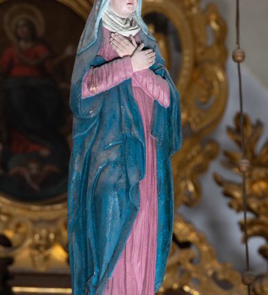 Zdjęcie nr 3: Rzeźba drewniana, pełnoplastyczna, polichromowana przedstawia Matkę Boską stojącą na niewielkim postumencie w kształcie zbliżonym do połowy sfery. Matka Boska ukazana w pozycji stojącej, frontalnie, w lekkim kontrapoście, z ciałem lekko zwróconym w kierunku krzyża, z rękoma skrzyżowanymi na piersi. Jej głowa jest nakryta welonem. Twarz ma owalną, oczy z opadającymi kącikami, nos prosty, a usta małe. Maria ubrana w długą suknię w kolorze zgaszonego różu przepasaną w stanie oraz niebieski płaszcz narzucony na ramiona  i opływający sylwetkę, na szyi udrapowana chusta. Fałdy szaty pionowe, płytkie, układające się miękko.