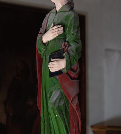 Zdjęcie nr 5: Rzeźba drewniana, pełnoplastyczna, polichromowana przedstawia stojącego na niewielkim postumencie św. Jana. Święty jest ukazany w pozycji stojącej, frontalnie, z lewą nogą ustawioną na niewielkim i wysuniętą do przodu, z ciałem lekko zwróconym w kierunku krzyża, z prawą ręką na piersi, z księgą w lewej dłoni. Jego głowa jest uniesiona, zwrócona ku Chrystusowi. Twarz owalna, oczy z opadającymi kącikami, nos prosty, usta małe. Włosy ciemne ułożone w pukle. Święty ubrany w długą, przepasaną w stanie zieloną suknię z kołnierzem oraz czerwono-brązowy płaszcz narzucony na prawe ramię i podtrzymywany ramieniem przy lewym boku. Fałdy szaty długie pionowe, układające się miękko.