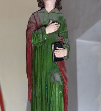 Zdjęcie nr 3: Rzeźba drewniana, pełnoplastyczna, polichromowana przedstawia stojącego na niewielkim postumencie św. Jana. Święty jest ukazany w pozycji stojącej, frontalnie, z lewą nogą ustawioną na niewielkim i wysuniętą do przodu, z ciałem lekko zwróconym w kierunku krzyża, z prawą ręką na piersi, z księgą w lewej dłoni. Jego głowa jest uniesiona, zwrócona ku Chrystusowi. Twarz owalna, oczy z opadającymi kącikami, nos prosty, usta małe. Włosy ciemne ułożone w pukle. Święty ubrany w długą, przepasaną w stanie zieloną suknię z kołnierzem oraz czerwono-brązowy płaszcz narzucony na prawe ramię i podtrzymywany ramieniem przy lewym boku. Fałdy szaty długie pionowe, układające się miękko.