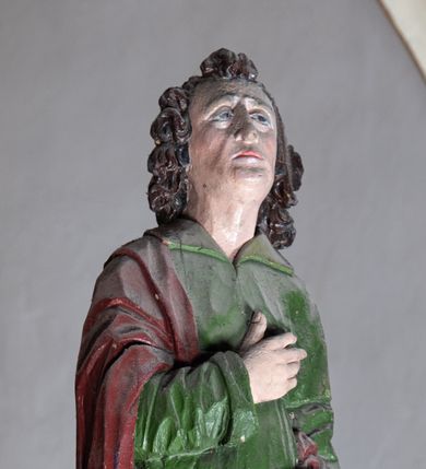 Zdjęcie nr 1: Rzeźba drewniana, pełnoplastyczna, polichromowana przedstawia stojącego na niewielkim postumencie św. Jana. Święty jest ukazany w pozycji stojącej, frontalnie, z lewą nogą ustawioną na niewielkim i wysuniętą do przodu, z ciałem lekko zwróconym w kierunku krzyża, z prawą ręką na piersi, z księgą w lewej dłoni. Jego głowa jest uniesiona, zwrócona ku Chrystusowi. Twarz owalna, oczy z opadającymi kącikami, nos prosty, usta małe. Włosy ciemne ułożone w pukle. Święty ubrany w długą, przepasaną w stanie zieloną suknię z kołnierzem oraz czerwono-brązowy płaszcz narzucony na prawe ramię i podtrzymywany ramieniem przy lewym boku. Fałdy szaty długie pionowe, układające się miękko.