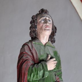 Zdjęcie nr 1: Rzeźba drewniana, pełnoplastyczna, polichromowana przedstawia stojącego na niewielkim postumencie św. Jana. Święty jest ukazany w pozycji stojącej, frontalnie, z lewą nogą ustawioną na niewielkim i wysuniętą do przodu, z ciałem lekko zwróconym w kierunku krzyża, z prawą ręką na piersi, z księgą w lewej dłoni. Jego głowa jest uniesiona, zwrócona ku Chrystusowi. Twarz owalna, oczy z opadającymi kącikami, nos prosty, usta małe. Włosy ciemne ułożone w pukle. Święty ubrany w długą, przepasaną w stanie zieloną suknię z kołnierzem oraz czerwono-brązowy płaszcz narzucony na prawe ramię i podtrzymywany ramieniem przy lewym boku. Fałdy szaty długie pionowe, układające się miękko.