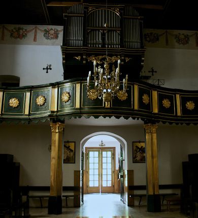 Zdjęcie nr 1: Chór muzyczny na zachodniej ścianie nawy, wsparty na dwóch słupach, ozdobionych od strony nawy dwoma kanelowanymi pilastrami o kapitelach kompozytowych. Jego parapet jest wklęsło-wypukły, z profilowanym cokołem, podzielony na kwatery kanelowanymi i złoconymi pilastrami. W kwaterach wypukłe pola kwadratowe o ćwierkolistych narożach ozdobione rzeźbionymi stylizowanymi rozetami. Parapet zwieńczony niewielkim gzymsem przełamanym na osi pilastrów. Pod listwą cokołu falisty fartuch. Struktura chóru muzycznego ciemnozielona. Pola płycin oliwkowo-ciemnozielone. Pilasty, rozety i profilowania są złocone. 
Znajdujący się na chórze prospekt wsparty na cokole z czterema kroksztynami, trzyosiowy, z osiami wydzielonymi kwadratowymi w podstawie filarkami. Oś środkowa podzielona na wie strefy: dolną z dwiema arkadkami z kostkowanym gzymsem i górną zamkniętą łukiem odcinkowym. Osie boczne zakończone gzymsem z kostkowaniem. W partii cokołowej bogato rzeźbiony motyw małżowinowo-chrząstkowy. 