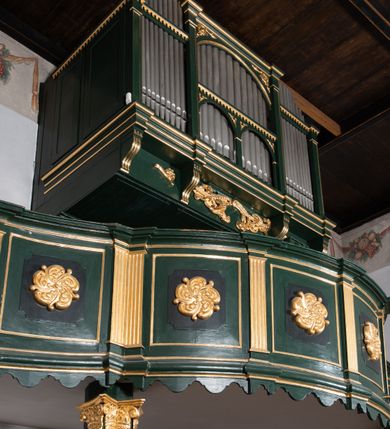 Zdjęcie nr 6: Chór muzyczny na zachodniej ścianie nawy, wsparty na dwóch słupach, ozdobionych od strony nawy dwoma kanelowanymi pilastrami o kapitelach kompozytowych. Jego parapet jest wklęsło-wypukły, z profilowanym cokołem, podzielony na kwatery kanelowanymi i złoconymi pilastrami. W kwaterach wypukłe pola kwadratowe o ćwierkolistych narożach ozdobione rzeźbionymi stylizowanymi rozetami. Parapet zwieńczony niewielkim gzymsem przełamanym na osi pilastrów. Pod listwą cokołu falisty fartuch. Struktura chóru muzycznego ciemnozielona. Pola płycin oliwkowo-ciemnozielone. Pilasty, rozety i profilowania są złocone. 
Znajdujący się na chórze prospekt wsparty na cokole z czterema kroksztynami, trzyosiowy, z osiami wydzielonymi kwadratowymi w podstawie filarkami. Oś środkowa podzielona na wie strefy: dolną z dwiema arkadkami z kostkowanym gzymsem i górną zamkniętą łukiem odcinkowym. Osie boczne zakończone gzymsem z kostkowaniem. W partii cokołowej bogato rzeźbiony motyw małżowinowo-chrząstkowy. 