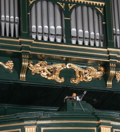 Zdjęcie nr 10: Chór muzyczny na zachodniej ścianie nawy, wsparty na dwóch słupach, ozdobionych od strony nawy dwoma kanelowanymi pilastrami o kapitelach kompozytowych. Jego parapet jest wklęsło-wypukły, z profilowanym cokołem, podzielony na kwatery kanelowanymi i złoconymi pilastrami. W kwaterach wypukłe pola kwadratowe o ćwierkolistych narożach ozdobione rzeźbionymi stylizowanymi rozetami. Parapet zwieńczony niewielkim gzymsem przełamanym na osi pilastrów. Pod listwą cokołu falisty fartuch. Struktura chóru muzycznego ciemnozielona. Pola płycin oliwkowo-ciemnozielone. Pilasty, rozety i profilowania są złocone. 
Znajdujący się na chórze prospekt wsparty na cokole z czterema kroksztynami, trzyosiowy, z osiami wydzielonymi kwadratowymi w podstawie filarkami. Oś środkowa podzielona na wie strefy: dolną z dwiema arkadkami z kostkowanym gzymsem i górną zamkniętą łukiem odcinkowym. Osie boczne zakończone gzymsem z kostkowaniem. W partii cokołowej bogato rzeźbiony motyw małżowinowo-chrząstkowy. 