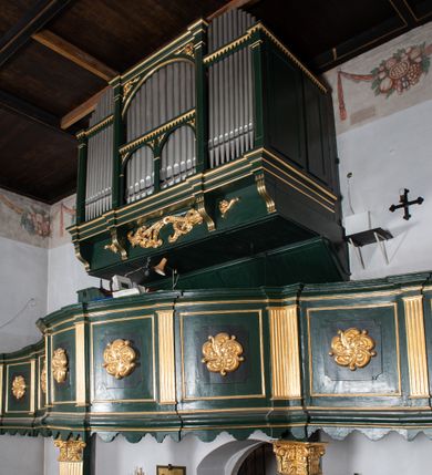 Zdjęcie nr 15: Chór muzyczny na zachodniej ścianie nawy, wsparty na dwóch słupach, ozdobionych od strony nawy dwoma kanelowanymi pilastrami o kapitelach kompozytowych. Jego parapet jest wklęsło-wypukły, z profilowanym cokołem, podzielony na kwatery kanelowanymi i złoconymi pilastrami. W kwaterach wypukłe pola kwadratowe o ćwierkolistych narożach ozdobione rzeźbionymi stylizowanymi rozetami. Parapet zwieńczony niewielkim gzymsem przełamanym na osi pilastrów. Pod listwą cokołu falisty fartuch. Struktura chóru muzycznego ciemnozielona. Pola płycin oliwkowo-ciemnozielone. Pilasty, rozety i profilowania są złocone. 
Znajdujący się na chórze prospekt wsparty na cokole z czterema kroksztynami, trzyosiowy, z osiami wydzielonymi kwadratowymi w podstawie filarkami. Oś środkowa podzielona na wie strefy: dolną z dwiema arkadkami z kostkowanym gzymsem i górną zamkniętą łukiem odcinkowym. Osie boczne zakończone gzymsem z kostkowaniem. W partii cokołowej bogato rzeźbiony motyw małżowinowo-chrząstkowy. 