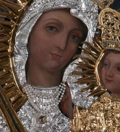 Zdjęcie nr 3: Obraz w kształcie prostokąta zamkniętego łukiem półpełnym, ujęty w profilowaną, złoconą ramą zdobioną kimationem. Tło niebiesko-szare, ze złotą, regencyjną kratą dekorowaną srebrnymi rozetami na przecięciach listew. Matka Boska ukazana w półpostaci, z głową zwróconą en trois quarts w lewo, prawą ręką uniesioną na wysokość piersi, podtrzymuje złote, tralkowate berło, lewą unosi Dzieciątko. Twarz owalna o beżowobrązowej karnacji, z prostym nosem, jasnoczerwonymi, uśmięchniętymi ustami, brązowymi oczami i regularnymi łukami brwiowymi. Głowa ujęta promienistą glorią. Chrystus siedzący na ramieniu matki, lewą ręką podtrzymuje zamkniętą księgę, prawą unosi na wysokość piersi w geście błogosławieństwa. Twarz, okrągła, o brązowobeżowej karnacji, z prostym nosem, lekko uśmiechniętymi ustami, brązowymi oczami, okolona krótkimi, prostymi włosami. Twarze i dłonie obu postaci odsłonięte, reszta przesłonięta srebrną sukienką z ornamentem roślinnym. Na głowie Matki Boskiej i Dzieciątka złocone, zamknięte, kameryzowane korony, zwieńczone krzyżami na kulach.