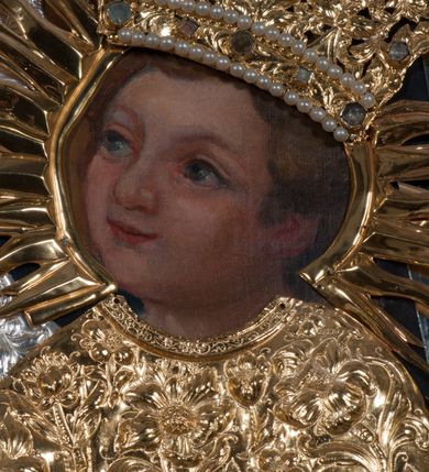 Zdjęcie nr 8: Obraz w kształcie prostokąta zamkniętego łukiem półpełnym, ujęty w profilowaną, złoconą ramą zdobioną kimationem. Tło niebiesko-szare, ze złotą, regencyjną kratą dekorowaną srebrnymi rozetami na przecięciach listew. Matka Boska ukazana w półpostaci, z głową zwróconą en trois quarts w lewo, prawą ręką uniesioną na wysokość piersi, podtrzymuje złote, tralkowate berło, lewą unosi Dzieciątko. Twarz owalna o beżowobrązowej karnacji, z prostym nosem, jasnoczerwonymi, uśmięchniętymi ustami, brązowymi oczami i regularnymi łukami brwiowymi. Głowa ujęta promienistą glorią. Chrystus siedzący na ramieniu matki, lewą ręką podtrzymuje zamkniętą księgę, prawą unosi na wysokość piersi w geście błogosławieństwa. Twarz, okrągła, o brązowobeżowej karnacji, z prostym nosem, lekko uśmiechniętymi ustami, brązowymi oczami, okolona krótkimi, prostymi włosami. Twarze i dłonie obu postaci odsłonięte, reszta przesłonięta srebrną sukienką z ornamentem roślinnym. Na głowie Matki Boskiej i Dzieciątka złocone, zamknięte, kameryzowane korony, zwieńczone krzyżami na kulach.