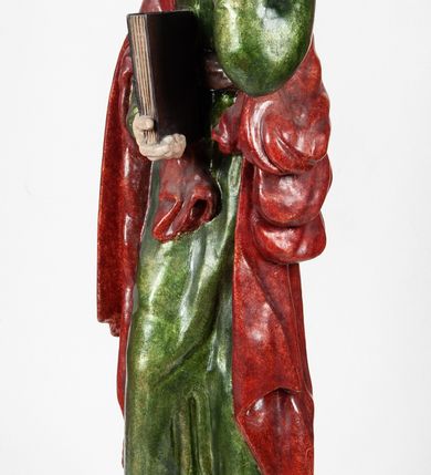 Zdjęcie nr 6: Rzeźba pełnoplastyczna, przedstawiająca św. Jana Ewangelistę w całej postaci. Apostoł stoi w lekkim kontrapoście, ciężar ciała wspierając na lewej nodze, z głową przechyloną na lewy bark. Prawą dłonią, uniesioną na wysokość pasa, podtrzymuje zamkniętą księgę, lewą dłoń przyciska do policzka w geście bólu i rozpaczy. Twarz potraktowana schematycznie, okolona długimi, brązowymi włosami z przedziałkiem pośrodku głowy. Święty ubrany w zieloną, długa tunikę, przewiązaną w pasie sznurem oraz czerwony płaszcz zarzucony na prawy bark i okrywający plecy, z przodu podtrzymywany prawym przedramieniem. Stopy bose.