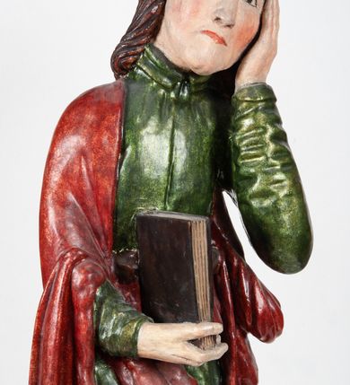 Zdjęcie nr 3: Rzeźba pełnoplastyczna, przedstawiająca św. Jana Ewangelistę w całej postaci. Apostoł stoi w lekkim kontrapoście, ciężar ciała wspierając na lewej nodze, z głową przechyloną na lewy bark. Prawą dłonią, uniesioną na wysokość pasa, podtrzymuje zamkniętą księgę, lewą dłoń przyciska do policzka w geście bólu i rozpaczy. Twarz potraktowana schematycznie, okolona długimi, brązowymi włosami z przedziałkiem pośrodku głowy. Święty ubrany w zieloną, długa tunikę, przewiązaną w pasie sznurem oraz czerwony płaszcz zarzucony na prawy bark i okrywający plecy, z przodu podtrzymywany prawym przedramieniem. Stopy bose.