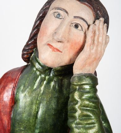 Zdjęcie nr 5: Rzeźba pełnoplastyczna, przedstawiająca św. Jana Ewangelistę w całej postaci. Apostoł stoi w lekkim kontrapoście, ciężar ciała wspierając na lewej nodze, z głową przechyloną na lewy bark. Prawą dłonią, uniesioną na wysokość pasa, podtrzymuje zamkniętą księgę, lewą dłoń przyciska do policzka w geście bólu i rozpaczy. Twarz potraktowana schematycznie, okolona długimi, brązowymi włosami z przedziałkiem pośrodku głowy. Święty ubrany w zieloną, długa tunikę, przewiązaną w pasie sznurem oraz czerwony płaszcz zarzucony na prawy bark i okrywający plecy, z przodu podtrzymywany prawym przedramieniem. Stopy bose.