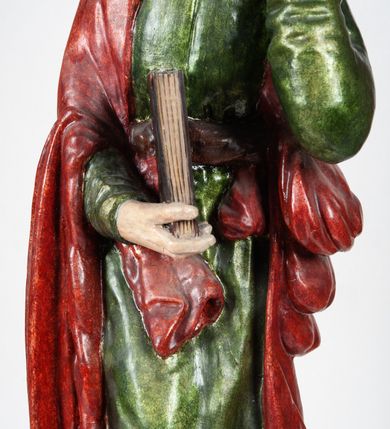 Zdjęcie nr 7: Rzeźba pełnoplastyczna, przedstawiająca św. Jana Ewangelistę w całej postaci. Apostoł stoi w lekkim kontrapoście, ciężar ciała wspierając na lewej nodze, z głową przechyloną na lewy bark. Prawą dłonią, uniesioną na wysokość pasa, podtrzymuje zamkniętą księgę, lewą dłoń przyciska do policzka w geście bólu i rozpaczy. Twarz potraktowana schematycznie, okolona długimi, brązowymi włosami z przedziałkiem pośrodku głowy. Święty ubrany w zieloną, długa tunikę, przewiązaną w pasie sznurem oraz czerwony płaszcz zarzucony na prawy bark i okrywający plecy, z przodu podtrzymywany prawym przedramieniem. Stopy bose.