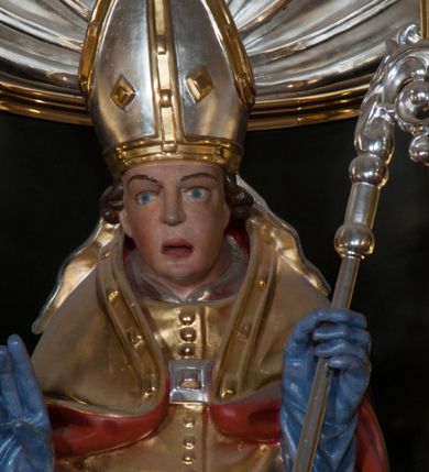 Zdjęcie nr 9: Rzeźba pełnoplastyczna, przeznaczona do umieszczenia w niszy, przedstawienie świętego w całej postaci umieszczone na niskim postumencie. Św. Stanisław ukazany jest frontalnie, w pozycji stojącej, w kontrapoście, z głową lekko pochyloną do przodu. Ręce ugięte w łokciach - w lewej pastorał, prawą wykonuje gest błogosławieństwa. Twarz pociągła, łukowato wygięte brwi, oczy szeroko otwarte, nos prosty, usta lekko rozchylone. Na głowie srebrno-złota infuła, spod której widoczne pukle kręconych włosów. Święty odziany jest w złotą kapę z czerwoną podszewką, spiętą na piersi fibulą, złoty mucet, rokietę, złotą albę, błękitne rękawiczki i brązowe buty. Rokieta fałdowana głęboko ciętymi, cienkimi, pionowymi fałdami, z jedną łukowato wygiętą fałdą. Alba z fałdami na kształt cienkich rurek, załamujących się u podstawy. Przy podstawie niewielka postać powstającego z grobu Piotrowina. Mężczyzna ukazany w trzech czwartych, zwrócony w lewo, z głową uniesioną w stronę świętego, prawą ręką na piersi, lewą wyprostowaną. Nogi ugięte w kolanach. Twarz pociągłą z wąsami. Włosy brązowe, krótkie, kręcone. Przepasany białą tkaniną, z ukośnymi, głęboko ciętymi fałdami.