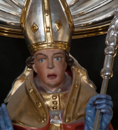 Zdjęcie nr 6: Rzeźba pełnoplastyczna, przeznaczona do umieszczenia w niszy, przedstawienie świętego w całej postaci umieszczone na niskim postumencie. Św. Stanisław ukazany jest frontalnie, w pozycji stojącej, w kontrapoście, z głową lekko pochyloną do przodu. Ręce ugięte w łokciach - w lewej pastorał, prawą wykonuje gest błogosławieństwa. Twarz pociągła, łukowato wygięte brwi, oczy szeroko otwarte, nos prosty, usta lekko rozchylone. Na głowie srebrno-złota infuła, spod której widoczne pukle kręconych włosów. Święty odziany jest w złotą kapę z czerwoną podszewką, spiętą na piersi fibulą, złoty mucet, rokietę, złotą albę, błękitne rękawiczki i brązowe buty. Rokieta fałdowana głęboko ciętymi, cienkimi, pionowymi fałdami, z jedną łukowato wygiętą fałdą. Alba z fałdami na kształt cienkich rurek, załamujących się u podstawy. Przy podstawie niewielka postać powstającego z grobu Piotrowina. Mężczyzna ukazany w trzech czwartych, zwrócony w lewo, z głową uniesioną w stronę świętego, prawą ręką na piersi, lewą wyprostowaną. Nogi ugięte w kolanach. Twarz pociągłą z wąsami. Włosy brązowe, krótkie, kręcone. Przepasany białą tkaniną, z ukośnymi, głęboko ciętymi fałdami.