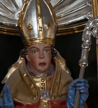 Zdjęcie nr 8: Rzeźba pełnoplastyczna, przeznaczona do umieszczenia w niszy, przedstawienie świętego w całej postaci umieszczone na niskim postumencie. Św. Stanisław ukazany jest frontalnie, w pozycji stojącej, w kontrapoście, z głową lekko pochyloną do przodu. Ręce ugięte w łokciach - w lewej pastorał, prawą wykonuje gest błogosławieństwa. Twarz pociągła, łukowato wygięte brwi, oczy szeroko otwarte, nos prosty, usta lekko rozchylone. Na głowie srebrno-złota infuła, spod której widoczne pukle kręconych włosów. Święty odziany jest w złotą kapę z czerwoną podszewką, spiętą na piersi fibulą, złoty mucet, rokietę, złotą albę, błękitne rękawiczki i brązowe buty. Rokieta fałdowana głęboko ciętymi, cienkimi, pionowymi fałdami, z jedną łukowato wygiętą fałdą. Alba z fałdami na kształt cienkich rurek, załamujących się u podstawy. Przy podstawie niewielka postać powstającego z grobu Piotrowina. Mężczyzna ukazany w trzech czwartych, zwrócony w lewo, z głową uniesioną w stronę świętego, prawą ręką na piersi, lewą wyprostowaną. Nogi ugięte w kolanach. Twarz pociągłą z wąsami. Włosy brązowe, krótkie, kręcone. Przepasany białą tkaniną, z ukośnymi, głęboko ciętymi fałdami.