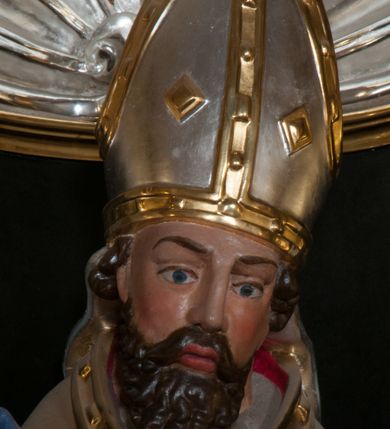 Zdjęcie nr 6: Rzeźba pełnoplastyczna, przyścienna, przeznaczona do umieszczenia w niszy, przedstawiająca św. Wojciecha w całej postaci, na niskim postumencie. Święty ukazany frontalnie, w pozycji stojącej, w kontrapoście, z głową lekko pochyloną. Ręce ugięte w łokciach, prawą dłonią trzyma wiosło. Pociągłą twarz okala ciemna, falowana broda i wąsy. Brwi cienkie, nos prosty. Na głowie srebrno-złota infuła, spod której widoczne pukle kręconych włosów. Święty odziany jest w złotą kapę z czerwoną podszewką, biały paliusz ze złotymi krzyżami, rokietę, złotą albę, błękitne rękawiczki i brązowe buty. Rokieta fałdowana głęboko ciętymi, cienkimi, pionowymi fałdami, z jedną łukowato wygiętą fałdą. Alba z fałdami na kształt cienkich rurek, załamujących się u podstawy.