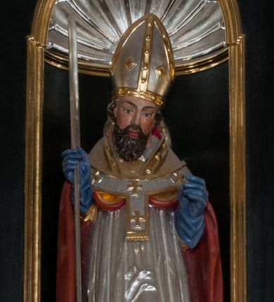 Zdjęcie nr 1: Rzeźba pełnoplastyczna, przyścienna, przeznaczona do umieszczenia w niszy, przedstawiająca św. Wojciecha w całej postaci, na niskim postumencie. Święty ukazany frontalnie, w pozycji stojącej, w kontrapoście, z głową lekko pochyloną. Ręce ugięte w łokciach, prawą dłonią trzyma wiosło. Pociągłą twarz okala ciemna, falowana broda i wąsy. Brwi cienkie, nos prosty. Na głowie srebrno-złota infuła, spod której widoczne pukle kręconych włosów. Święty odziany jest w złotą kapę z czerwoną podszewką, biały paliusz ze złotymi krzyżami, rokietę, złotą albę, błękitne rękawiczki i brązowe buty. Rokieta fałdowana głęboko ciętymi, cienkimi, pionowymi fałdami, z jedną łukowato wygiętą fałdą. Alba z fałdami na kształt cienkich rurek, załamujących się u podstawy.