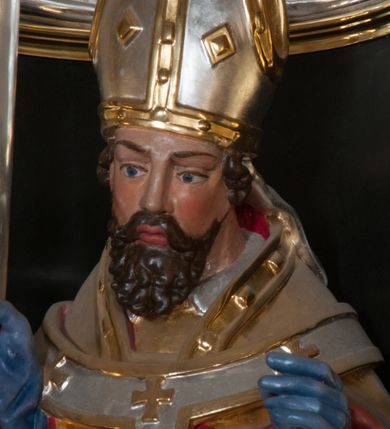 Zdjęcie nr 5: Rzeźba pełnoplastyczna, przyścienna, przeznaczona do umieszczenia w niszy, przedstawiająca św. Wojciecha w całej postaci, na niskim postumencie. Święty ukazany frontalnie, w pozycji stojącej, w kontrapoście, z głową lekko pochyloną. Ręce ugięte w łokciach, prawą dłonią trzyma wiosło. Pociągłą twarz okala ciemna, falowana broda i wąsy. Brwi cienkie, nos prosty. Na głowie srebrno-złota infuła, spod której widoczne pukle kręconych włosów. Święty odziany jest w złotą kapę z czerwoną podszewką, biały paliusz ze złotymi krzyżami, rokietę, złotą albę, błękitne rękawiczki i brązowe buty. Rokieta fałdowana głęboko ciętymi, cienkimi, pionowymi fałdami, z jedną łukowato wygiętą fałdą. Alba z fałdami na kształt cienkich rurek, załamujących się u podstawy.