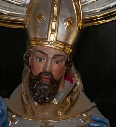 Zdjęcie nr 7: Rzeźba pełnoplastyczna, przyścienna, przeznaczona do umieszczenia w niszy, przedstawiająca św. Wojciecha w całej postaci, na niskim postumencie. Święty ukazany frontalnie, w pozycji stojącej, w kontrapoście, z głową lekko pochyloną. Ręce ugięte w łokciach, prawą dłonią trzyma wiosło. Pociągłą twarz okala ciemna, falowana broda i wąsy. Brwi cienkie, nos prosty. Na głowie srebrno-złota infuła, spod której widoczne pukle kręconych włosów. Święty odziany jest w złotą kapę z czerwoną podszewką, biały paliusz ze złotymi krzyżami, rokietę, złotą albę, błękitne rękawiczki i brązowe buty. Rokieta fałdowana głęboko ciętymi, cienkimi, pionowymi fałdami, z jedną łukowato wygiętą fałdą. Alba z fałdami na kształt cienkich rurek, załamujących się u podstawy.