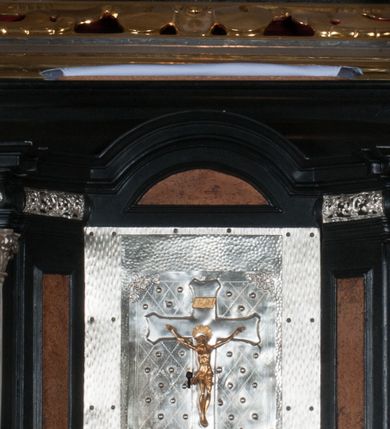 Zdjęcie nr 1: Tabernakulum na rzucie połowy ośmioboku, architektoniczne, na niskim postumencie, trójosiowe, dwudzielne (z wydzieloną gzymsem częścią cokołową), artykułowane kolumienkami o kapitelach kompozytowych oraz wolutami podtrzymującymi kompozytowe kapitele, na wybrzuszonych, zdwojonych cokołach. Belkowanie występujące nad podporami. Ścianka z drzwiczkami zamknięta łukiem półkolistym. Cokoliki, fryz i boczne ścianki tabernakulum zdobione suchym akantem. Drzwiczki jednoskrzydłowe (nowsze, pierwotne dwuskrzydłowe), srebrne, zdobione prostokątną płyciną, z romboidalnymi polami i spłaszczonymi kulami. W narożach stylizowany ornament roślinny. W centrum przedstawienie Chrystusa na krzyżu. Pola bocznych ścianek zdobione imitującymi szufladki mozaikowymi płycinami z różnobarwnych marmurów przedstawiającymi pejzaż z elementami architektury oraz płycinami wykładanymi jasnym drewnem. Pejzażowe płyciny w górnej części tabernakulum ujęte srebrnymi, odlewanymi i cyzelowanym ramkami. 
Na tabernakulum umieszczone antytetycznie rzeźby dwóch aniołów: pełnoplastyczne, w ujęciu całopostaciowym, wolnostojące. Anioł po lewej stronie ukazany jest w pozycji klęczącej, z profilu, pochylony do przodu. W rękach trzyma łódkę na kadzidło. Włosy brązowe, falowane, sięgające do ramion. Twarz pociągła, łukowato wygięte brwi, szeroko otwarte oczy, prosty nos, róż na policzkach. Skrzydła złote. Anioł odziany jest białą tunikę, z krótkimi rękawami i z cienkimi, pionowymi fałdami, załamującymi się na wysokości kolan. Stopy bose. Anioł po prawej ukazany jest w pozycji klęczącej, z profilu, pochylony do przodu. W rękach trzyma trybularz. Włosy brązowe, falowane, sięgające do ramion. Twarz pociągła, łukowato wygięte brwi, szeroko otwarte oczy, prosty nos, róż na policzkach. Skrzydła złote. Anioł odziany jest białą tunikę, z krótkimi rękawami i z cienkimi, pionowymi fałdami, załamującymi się na wysokości kolan. Stopy bose.