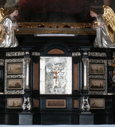 Zdjęcie nr 6: Tabernakulum na rzucie połowy ośmioboku, architektoniczne, na niskim postumencie, trójosiowe, dwudzielne (z wydzieloną gzymsem częścią cokołową), artykułowane kolumienkami o kapitelach kompozytowych oraz wolutami podtrzymującymi kompozytowe kapitele, na wybrzuszonych, zdwojonych cokołach. Belkowanie występujące nad podporami. Ścianka z drzwiczkami zamknięta łukiem półkolistym. Cokoliki, fryz i boczne ścianki tabernakulum zdobione suchym akantem. Drzwiczki jednoskrzydłowe (nowsze, pierwotne dwuskrzydłowe), srebrne, zdobione prostokątną płyciną, z romboidalnymi polami i spłaszczonymi kulami. W narożach stylizowany ornament roślinny. W centrum przedstawienie Chrystusa na krzyżu. Pola bocznych ścianek zdobione imitującymi szufladki mozaikowymi płycinami z różnobarwnych marmurów przedstawiającymi pejzaż z elementami architektury oraz płycinami wykładanymi jasnym drewnem. Pejzażowe płyciny w górnej części tabernakulum ujęte srebrnymi, odlewanymi i cyzelowanym ramkami. 
Na tabernakulum umieszczone antytetycznie rzeźby dwóch aniołów: pełnoplastyczne, w ujęciu całopostaciowym, wolnostojące. Anioł po lewej stronie ukazany jest w pozycji klęczącej, z profilu, pochylony do przodu. W rękach trzyma łódkę na kadzidło. Włosy brązowe, falowane, sięgające do ramion. Twarz pociągła, łukowato wygięte brwi, szeroko otwarte oczy, prosty nos, róż na policzkach. Skrzydła złote. Anioł odziany jest białą tunikę, z krótkimi rękawami i z cienkimi, pionowymi fałdami, załamującymi się na wysokości kolan. Stopy bose. Anioł po prawej ukazany jest w pozycji klęczącej, z profilu, pochylony do przodu. W rękach trzyma trybularz. Włosy brązowe, falowane, sięgające do ramion. Twarz pociągła, łukowato wygięte brwi, szeroko otwarte oczy, prosty nos, róż na policzkach. Skrzydła złote. Anioł odziany jest białą tunikę, z krótkimi rękawami i z cienkimi, pionowymi fałdami, załamującymi się na wysokości kolan. Stopy bose.