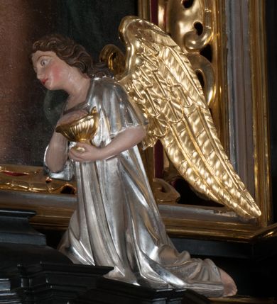 Zdjęcie nr 10: Tabernakulum na rzucie połowy ośmioboku, architektoniczne, na niskim postumencie, trójosiowe, dwudzielne (z wydzieloną gzymsem częścią cokołową), artykułowane kolumienkami o kapitelach kompozytowych oraz wolutami podtrzymującymi kompozytowe kapitele, na wybrzuszonych, zdwojonych cokołach. Belkowanie występujące nad podporami. Ścianka z drzwiczkami zamknięta łukiem półkolistym. Cokoliki, fryz i boczne ścianki tabernakulum zdobione suchym akantem. Drzwiczki jednoskrzydłowe (nowsze, pierwotne dwuskrzydłowe), srebrne, zdobione prostokątną płyciną, z romboidalnymi polami i spłaszczonymi kulami. W narożach stylizowany ornament roślinny. W centrum przedstawienie Chrystusa na krzyżu. Pola bocznych ścianek zdobione imitującymi szufladki mozaikowymi płycinami z różnobarwnych marmurów przedstawiającymi pejzaż z elementami architektury oraz płycinami wykładanymi jasnym drewnem. Pejzażowe płyciny w górnej części tabernakulum ujęte srebrnymi, odlewanymi i cyzelowanym ramkami. 
Na tabernakulum umieszczone antytetycznie rzeźby dwóch aniołów: pełnoplastyczne, w ujęciu całopostaciowym, wolnostojące. Anioł po lewej stronie ukazany jest w pozycji klęczącej, z profilu, pochylony do przodu. W rękach trzyma łódkę na kadzidło. Włosy brązowe, falowane, sięgające do ramion. Twarz pociągła, łukowato wygięte brwi, szeroko otwarte oczy, prosty nos, róż na policzkach. Skrzydła złote. Anioł odziany jest białą tunikę, z krótkimi rękawami i z cienkimi, pionowymi fałdami, załamującymi się na wysokości kolan. Stopy bose. Anioł po prawej ukazany jest w pozycji klęczącej, z profilu, pochylony do przodu. W rękach trzyma trybularz. Włosy brązowe, falowane, sięgające do ramion. Twarz pociągła, łukowato wygięte brwi, szeroko otwarte oczy, prosty nos, róż na policzkach. Skrzydła złote. Anioł odziany jest białą tunikę, z krótkimi rękawami i z cienkimi, pionowymi fałdami, załamującymi się na wysokości kolan. Stopy bose.