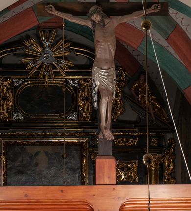 Zdjęcie nr 6: Chrystus wiszący na ramionach łagodnym łukiem rozpostartych na krzyżu, o smukłej sylwecie, z nogami skrzyżowanymi, lekko ugiętymi w kolanach. Ciało zmarłego Chrystusa ukazane jest realistycznie, z głową silnie przechyloną na prawe ramie, zamkniętymi oczami, długim nosem i otwartymi ustami. Twarz okala krótka, spiczasta broda i wąsy, włosy opadają na kark i klatkę piersiową, na głowie korona cierniowa w kształcie skręconego sznura z kolcami. Klatka piersiowa o owalnym obrysie, z zaznaczonymi żebrami. Stosunkowo długie perizonium, zdobione srebrzeniem na pulmencie, układa się z przodu łukowatymi fałdami i spływa z bioder dekoracyjnie traktowanymi zwisami. Na ciele widoczna rana od włóczni, z niej i z ran na rękach i nogach widoczne spływające strużki krwi. 
