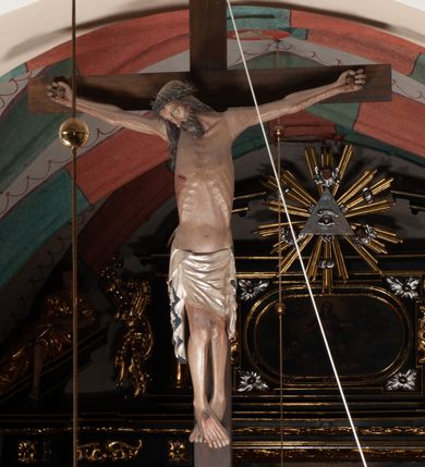 Zdjęcie nr 2: Chrystus wiszący na ramionach łagodnym łukiem rozpostartych na krzyżu, o smukłej sylwecie, z nogami skrzyżowanymi, lekko ugiętymi w kolanach. Ciało zmarłego Chrystusa ukazane jest realistycznie, z głową silnie przechyloną na prawe ramie, zamkniętymi oczami, długim nosem i otwartymi ustami. Twarz okala krótka, spiczasta broda i wąsy, włosy opadają na kark i klatkę piersiową, na głowie korona cierniowa w kształcie skręconego sznura z kolcami. Klatka piersiowa o owalnym obrysie, z zaznaczonymi żebrami. Stosunkowo długie perizonium, zdobione srebrzeniem na pulmencie, układa się z przodu łukowatymi fałdami i spływa z bioder dekoracyjnie traktowanymi zwisami. Na ciele widoczna rana od włóczni, z niej i z ran na rękach i nogach widoczne spływające strużki krwi. 
