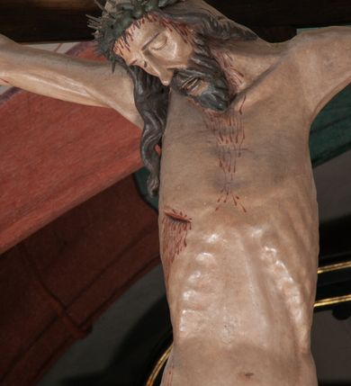 Zdjęcie nr 4: Chrystus wiszący na ramionach łagodnym łukiem rozpostartych na krzyżu, o smukłej sylwecie, z nogami skrzyżowanymi, lekko ugiętymi w kolanach. Ciało zmarłego Chrystusa ukazane jest realistycznie, z głową silnie przechyloną na prawe ramie, zamkniętymi oczami, długim nosem i otwartymi ustami. Twarz okala krótka, spiczasta broda i wąsy, włosy opadają na kark i klatkę piersiową, na głowie korona cierniowa w kształcie skręconego sznura z kolcami. Klatka piersiowa o owalnym obrysie, z zaznaczonymi żebrami. Stosunkowo długie perizonium, zdobione srebrzeniem na pulmencie, układa się z przodu łukowatymi fałdami i spływa z bioder dekoracyjnie traktowanymi zwisami. Na ciele widoczna rana od włóczni, z niej i z ran na rękach i nogach widoczne spływające strużki krwi. 
