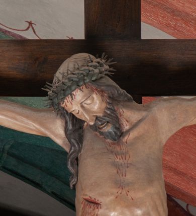 Zdjęcie nr 5: Chrystus wiszący na ramionach łagodnym łukiem rozpostartych na krzyżu, o smukłej sylwecie, z nogami skrzyżowanymi, lekko ugiętymi w kolanach. Ciało zmarłego Chrystusa ukazane jest realistycznie, z głową silnie przechyloną na prawe ramie, zamkniętymi oczami, długim nosem i otwartymi ustami. Twarz okala krótka, spiczasta broda i wąsy, włosy opadają na kark i klatkę piersiową, na głowie korona cierniowa w kształcie skręconego sznura z kolcami. Klatka piersiowa o owalnym obrysie, z zaznaczonymi żebrami. Stosunkowo długie perizonium, zdobione srebrzeniem na pulmencie, układa się z przodu łukowatymi fałdami i spływa z bioder dekoracyjnie traktowanymi zwisami. Na ciele widoczna rana od włóczni, z niej i z ran na rękach i nogach widoczne spływające strużki krwi. 
