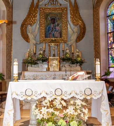 Zdjęcie nr 2: Nastawa niearchitektoniczna. Ołtarz prostopadłościenny, kamienny, z drewnianą mensą. Antepedium dekorowane trzema kwadratowymi polami wypełnionymi geometryczną mozaiką w formie krzyży o rozszerzających się ramionach. Na ołtarzu tabernakulum w formie prostopadłościennej skrzynki, obitej blachą, o zaokrąglonych narożach. Drzwiczki w kształcie stojącego prostokąta zamkniętego łukiem półkolistym nadwieszonym, z przedstawieniem kielicha z hostią w glorii. Na ściance frontowej aplikowana dekoracja w postaci kłosów zbóż i winnego grona. Tron wystawienia w formie baldachimu podtrzymywanego na czterech tralkach. Obraz Matki Boskiej Częstochowskiej w kształcie stojącego prostokąta, ujęty grubą, złoconą ramą dekorowaną ornamentem z lilii heraldycznych. Dzieło flankowane jest dwiema postaciami klęczących aniołów, podtrzymujących je obiema rękami. Anioły ukazane z profilu, lewy z głową skierowaną frontalnie. Ubrane są w luźne tuniki przepasane w talii. Skrzydła mają duże, złożone, wygięte na kształt litery „C”. Nad obrazem monogram Marii, flankowany przez dwa putta podtrzymujące nad nim koronę otwartą. Postacie polichromowane są na biało, o złoconych skrzydłach. 

