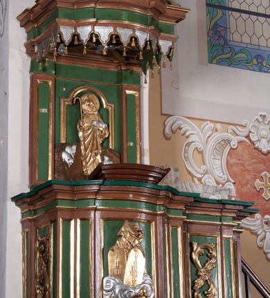 Zdjęcie nr 3: Ambona przyścienna składająca się z kosza, zaplecka, baldachimu i schodów z balustradą, zawieszona na lewej ścianie prezbiterium. Kosz na planie zbliżonym do prostokąta, o ściankach prostych i wklęsłych z uskokami w narożach, dekorowanymi płycinami z plakietami ornamentalnymi. Na największej płycinie przedstawienie Boga Ojca trzymającego tablice 10 przykazań, pośród obłoków, z trąbiącym aniołem w prawym dolnym narożniku. Kosz zamyka pełne belkowanie z silnie wysuniętym gzymsem. Podstawa kosza z łączących się pośrodku wolut, z pozłacaną szyszką u dołu. Zaplecek w kształcie prostokąta z prostokątną płyciną zamknięta łukiem nadwieszonym, wewnątrz której płaskorzeźbione przedstawienie Chrystusa Dobrego Pasterza. Pole flankują pilastry z płycinami. Baldachim powtarzający plan kosza, z podwieszonym lambrekinem i zwieńczony postumentem utworzonym ze stylizowanych wolut, na którym posadowiono figurę modlącego się anioła w długiej, srebrnej sukni. W podniebiu baldachimu znajduje się rzeźba srebrnej gołębicy Ducha Świętego na tle glorii promienistej. Struktura w kolorze brązowym, płyciny ciemnozielone, detal architektoniczny i ornamenty pozłacane i posrebrzane.
