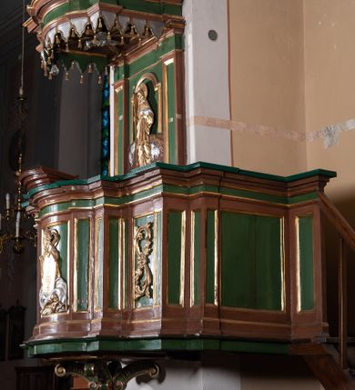 Zdjęcie nr 1: Ambona przyścienna składająca się z kosza, zaplecka, baldachimu i schodów z balustradą, zawieszona na lewej ścianie prezbiterium. Kosz na planie zbliżonym do prostokąta, o ściankach prostych i wklęsłych z uskokami w narożach, dekorowanymi płycinami z plakietami ornamentalnymi. Na największej płycinie przedstawienie Boga Ojca trzymającego tablice 10 przykazań, pośród obłoków, z trąbiącym aniołem w prawym dolnym narożniku. Kosz zamyka pełne belkowanie z silnie wysuniętym gzymsem. Podstawa kosza z łączących się pośrodku wolut, z pozłacaną szyszką u dołu. Zaplecek w kształcie prostokąta z prostokątną płyciną zamknięta łukiem nadwieszonym, wewnątrz której płaskorzeźbione przedstawienie Chrystusa Dobrego Pasterza. Pole flankują pilastry z płycinami. Baldachim powtarzający plan kosza, z podwieszonym lambrekinem i zwieńczony postumentem utworzonym ze stylizowanych wolut, na którym posadowiono figurę modlącego się anioła w długiej, srebrnej sukni. W podniebiu baldachimu znajduje się rzeźba srebrnej gołębicy Ducha Świętego na tle glorii promienistej. Struktura w kolorze brązowym, płyciny ciemnozielone, detal architektoniczny i ornamenty pozłacane i posrebrzane.
