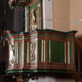 Zdjęcie nr 1: Ambona przyścienna składająca się z kosza, zaplecka, baldachimu i schodów z balustradą, zawieszona na lewej ścianie prezbiterium. Kosz na planie zbliżonym do prostokąta, o ściankach prostych i wklęsłych z uskokami w narożach, dekorowanymi płycinami z plakietami ornamentalnymi. Na największej płycinie przedstawienie Boga Ojca trzymającego tablice 10 przykazań, pośród obłoków, z trąbiącym aniołem w prawym dolnym narożniku. Kosz zamyka pełne belkowanie z silnie wysuniętym gzymsem. Podstawa kosza z łączących się pośrodku wolut, z pozłacaną szyszką u dołu. Zaplecek w kształcie prostokąta z prostokątną płyciną zamknięta łukiem nadwieszonym, wewnątrz której płaskorzeźbione przedstawienie Chrystusa Dobrego Pasterza. Pole flankują pilastry z płycinami. Baldachim powtarzający plan kosza, z podwieszonym lambrekinem i zwieńczony postumentem utworzonym ze stylizowanych wolut, na którym posadowiono figurę modlącego się anioła w długiej, srebrnej sukni. W podniebiu baldachimu znajduje się rzeźba srebrnej gołębicy Ducha Świętego na tle glorii promienistej. Struktura w kolorze brązowym, płyciny ciemnozielone, detal architektoniczny i ornamenty pozłacane i posrebrzane.
